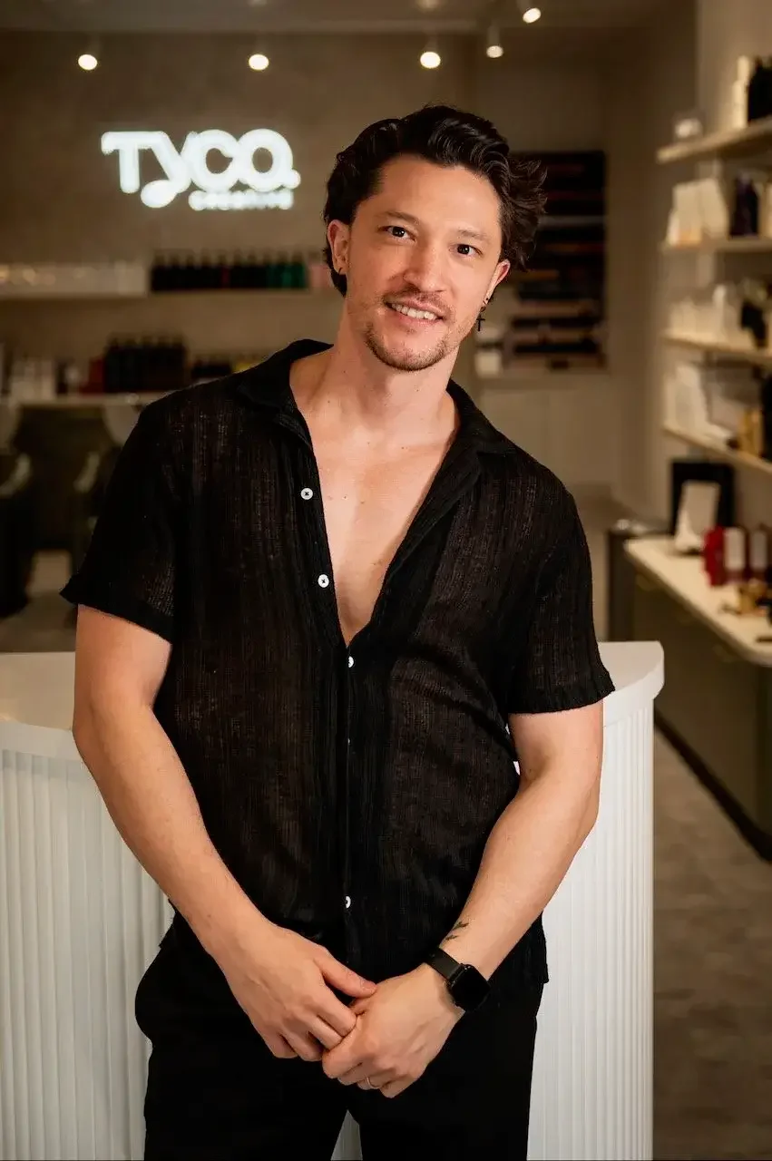 A man with dark, wavy hair and a light beard, wearing a partially unbuttoned black shirt, stands inside a modern coffee shop with a