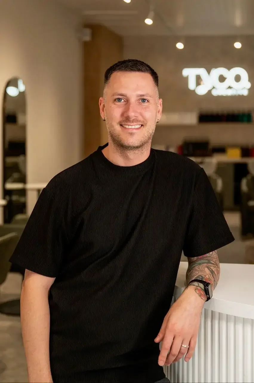 A smiling man with short dark hair, wearing a black t-shirt, standing in a modern office environment with a TYCO logo in the background.