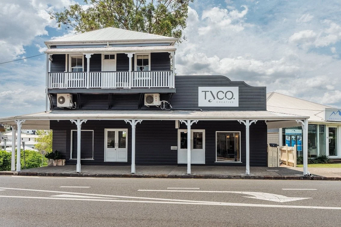 A two-story dark gray building with white trim, featuring a large sign that says TACO. Creative, located on a street with a parking lane and cloudy sky overhead.