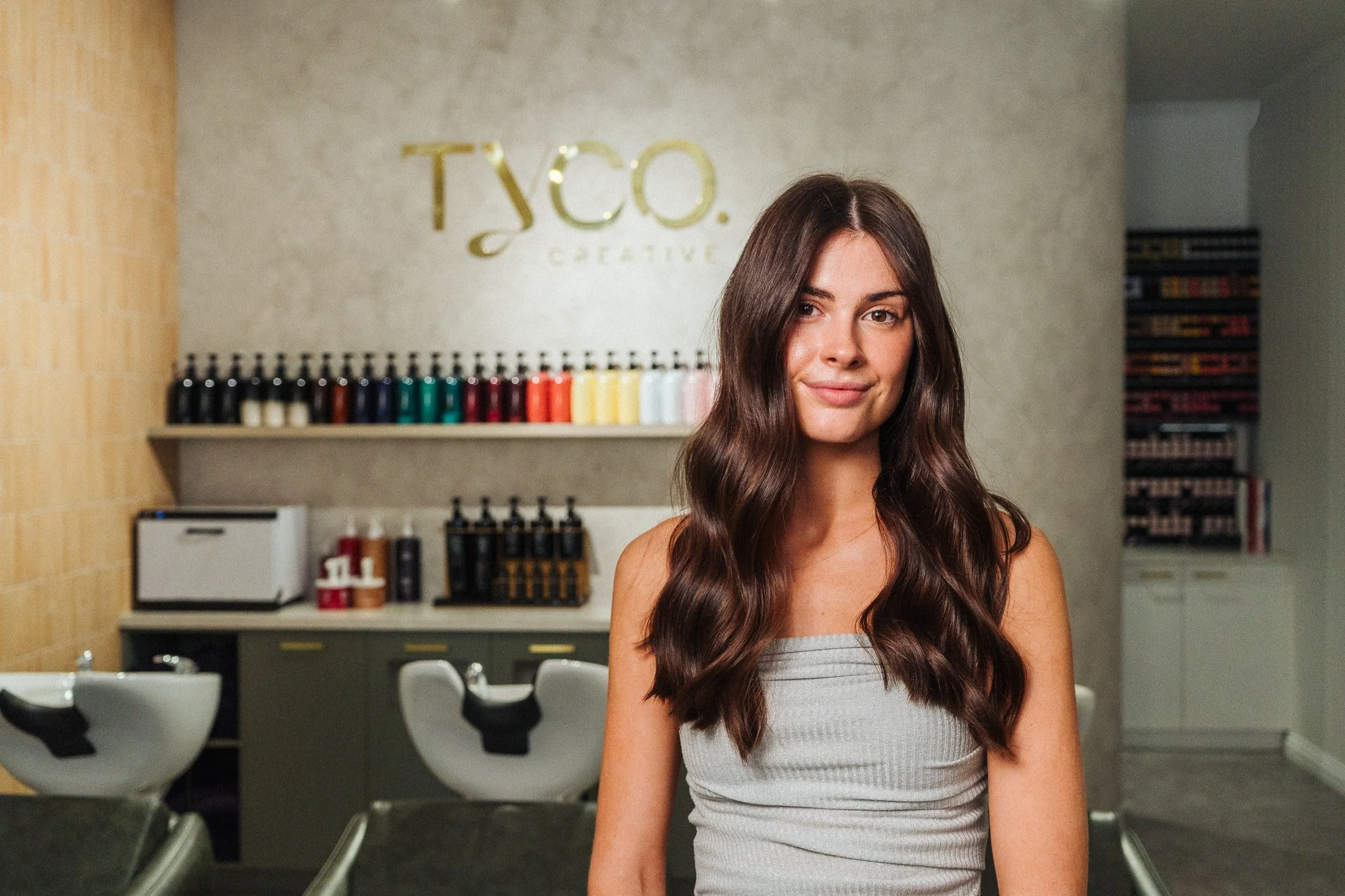 Young woman with long wavy brown hair smiling in salon with hair dye bottles on shelf and salon chairs in background.