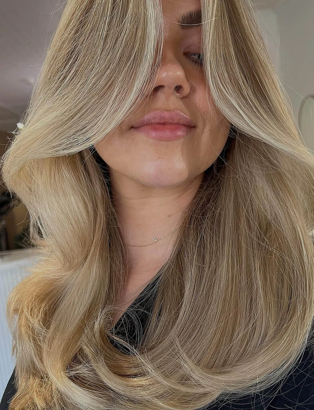Close-up of a woman with long, voluminous blonde hair covering part of her face, wearing makeup, pink lipstick, and a necklace with a small pendant.