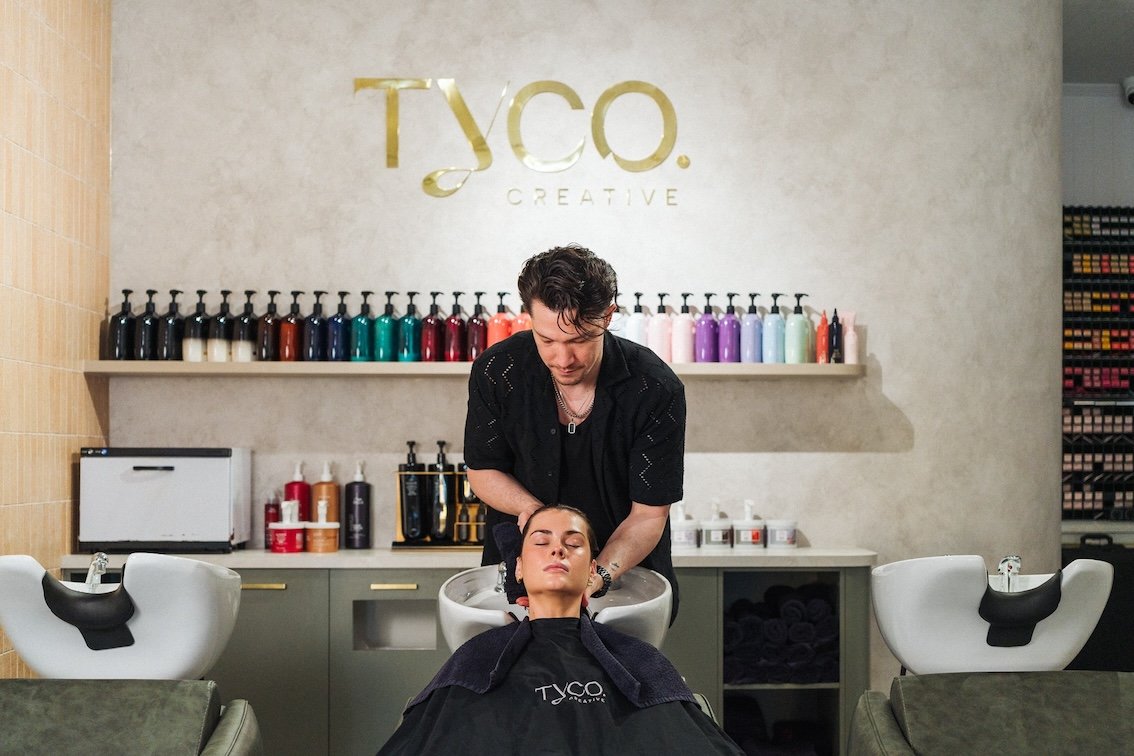 A woman with dark hair is getting her hair washed at a salon, with her eyes closed. A hairstylist is working on her hair, standing behind her. The salon has a wall with colorful bottles and a sign that says "TYCO CREATIVE." There are two washing stations with white sinks on either side of them.