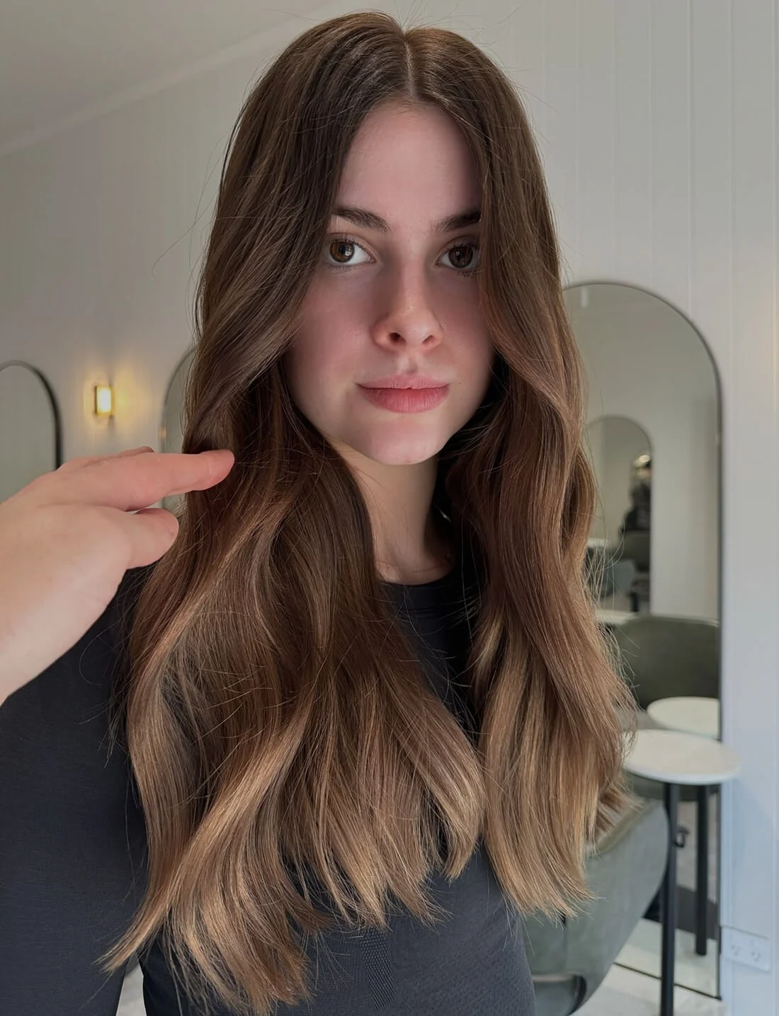 Close-up of a young woman with long brown wavy hair, wearing a black top, standing in a room with white walls, mirrors, and lighting fixtures.