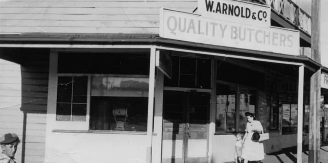 Black and white photo of a storefront with a sign that reads "W. Arnold & Co. Quality Butchers." A woman with a hat and a coat is walking with a child in front of the store.
