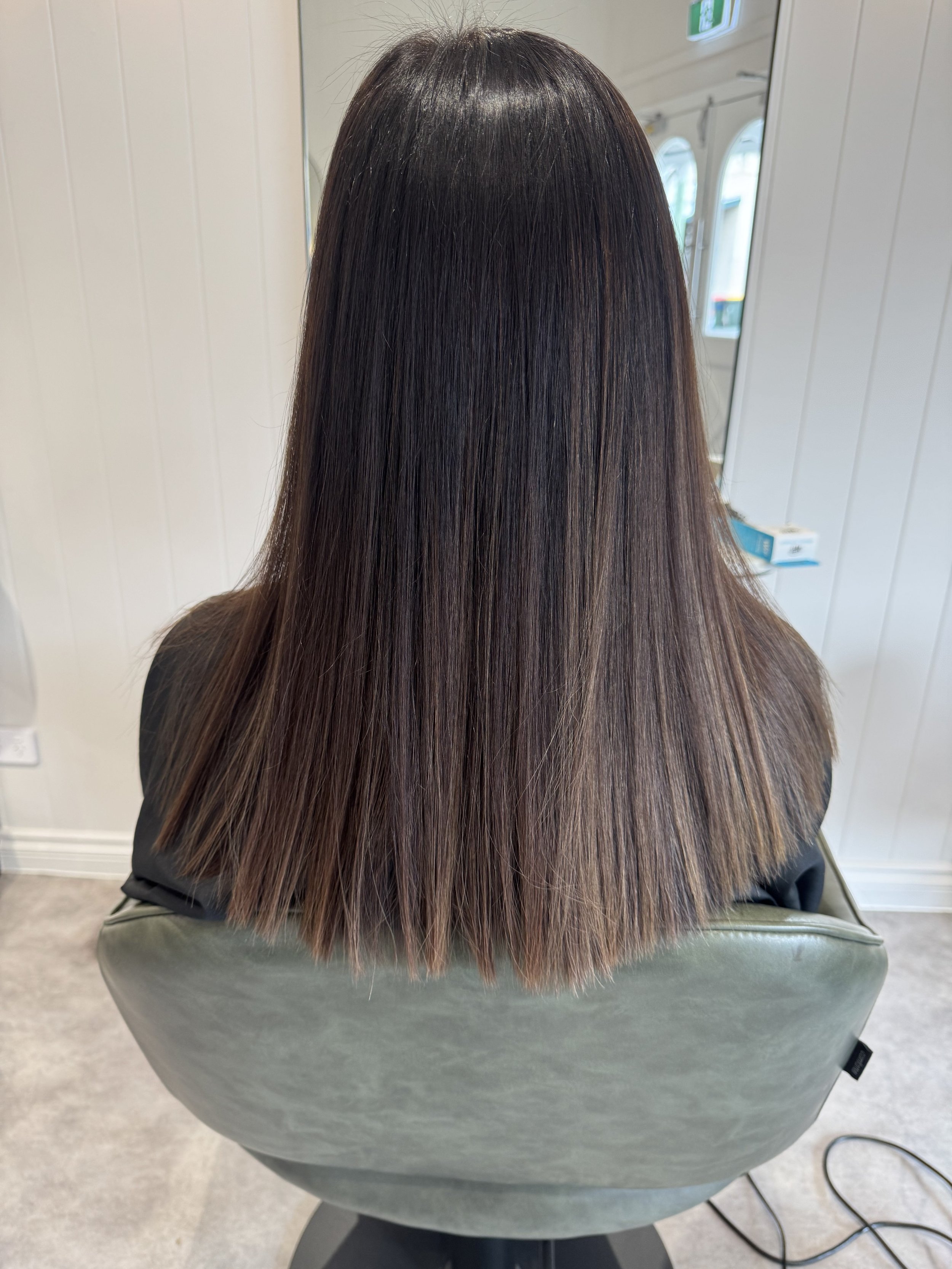 Back view of a woman with straight, shoulder-length brown hair sitting in a salon chair.