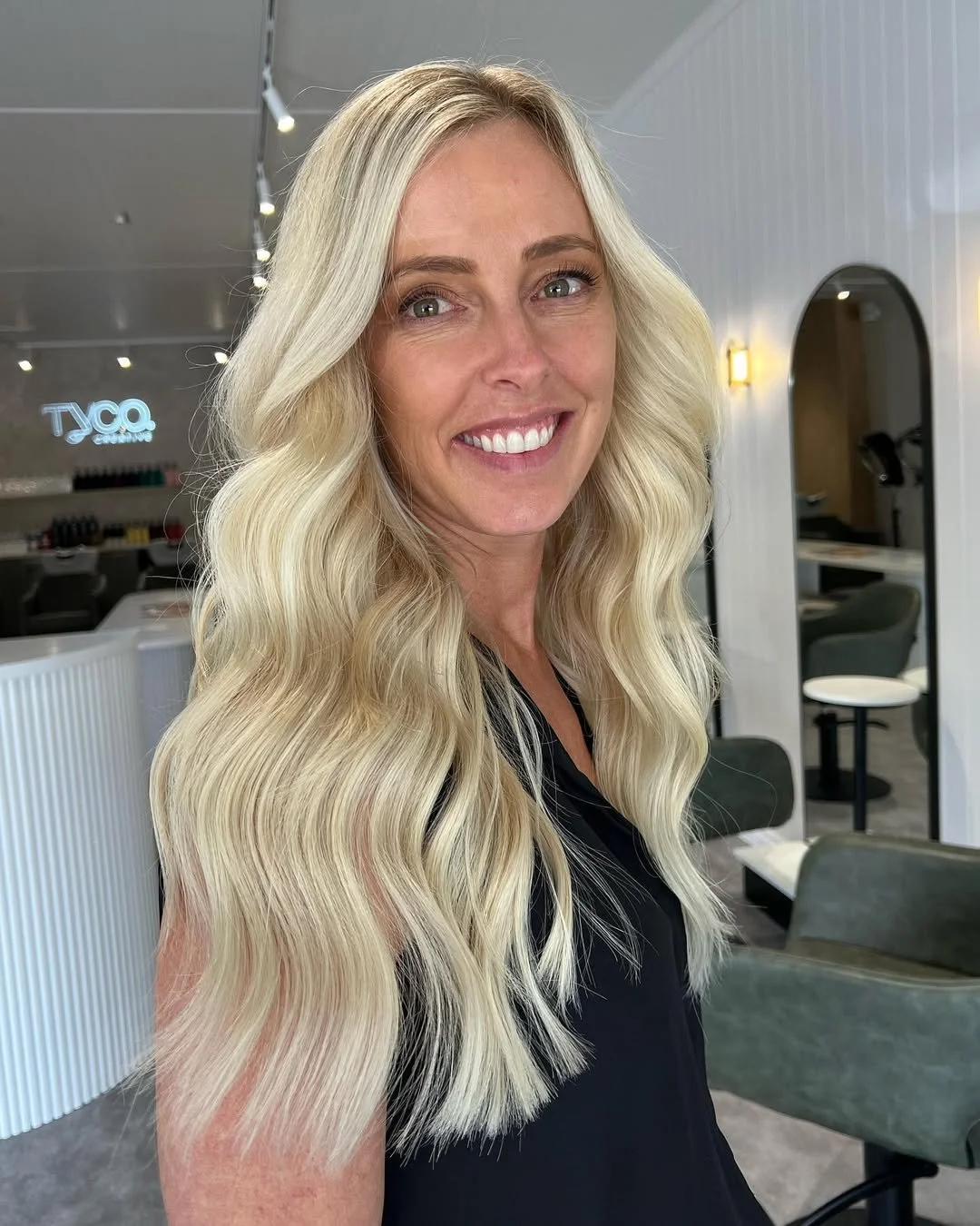 Woman with blonde hair and a slight smile, holding a lock of her hair near her face, wearing a white sleeveless top.