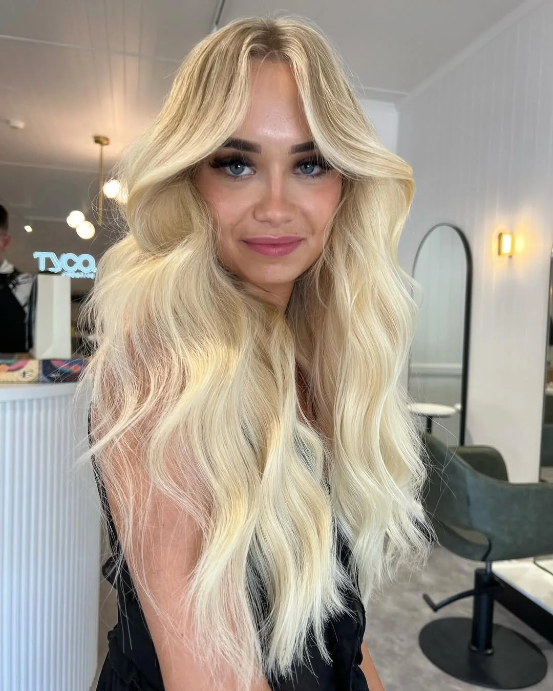 A woman with long, wavy, light brown hair and blue eyes smiling softly in a salon setting.