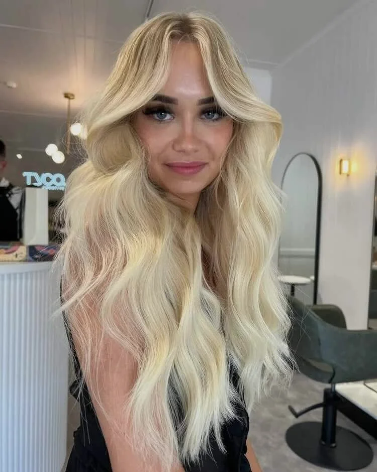 A woman with long, wavy blonde hair and bright blue eyes smiling indoors.