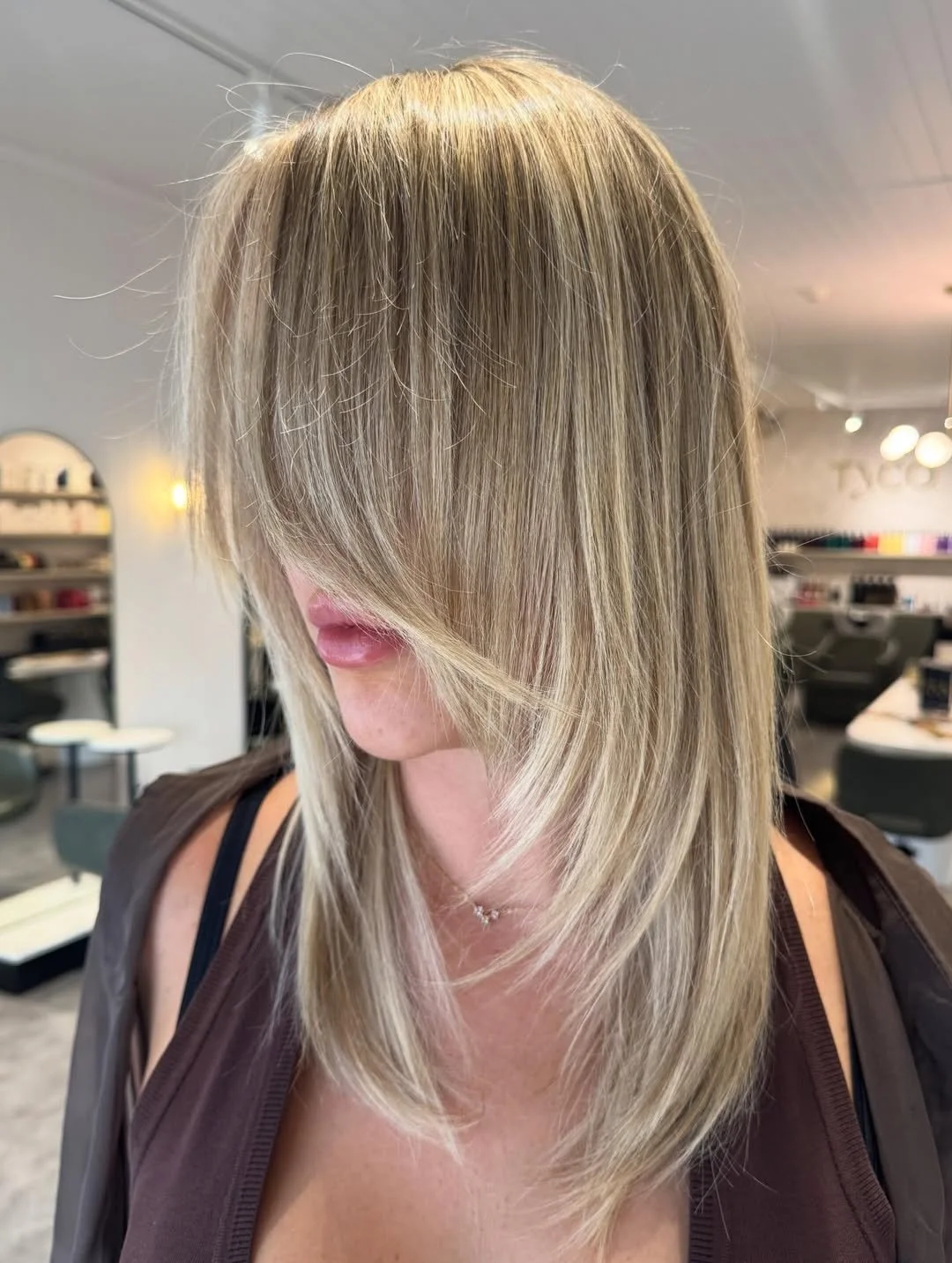 A young woman with long, wavy brown hair and a fair complexion looks into the camera. She is wearing a black top and is standing in front of a mirror with a minimalist interior background.