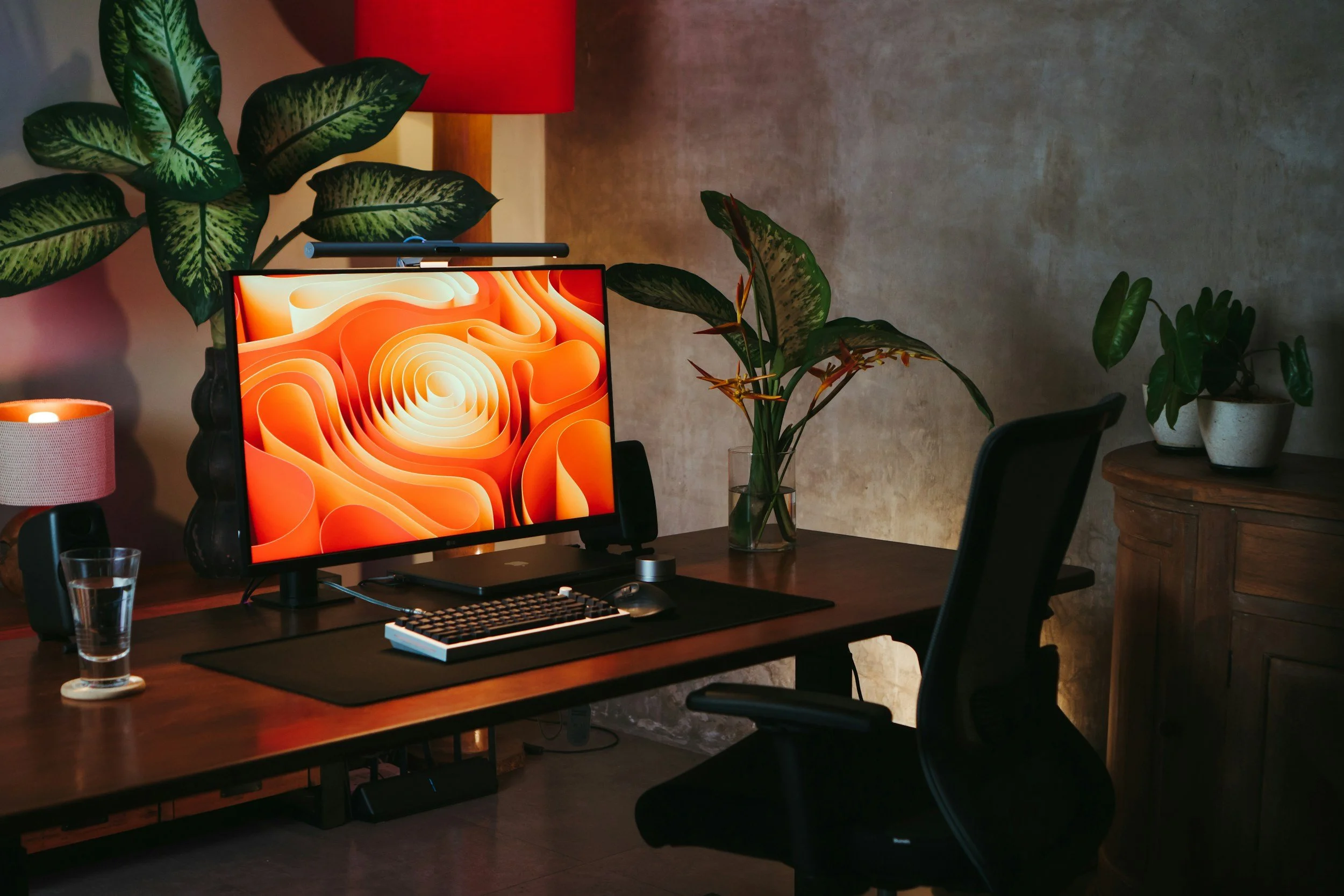 A home office desk with a computer monitor displaying an orange and cream abstract design, a keyboard, mouse, and a glass of water. There are potted plants, a red lampshade, and a small candle on the desk.