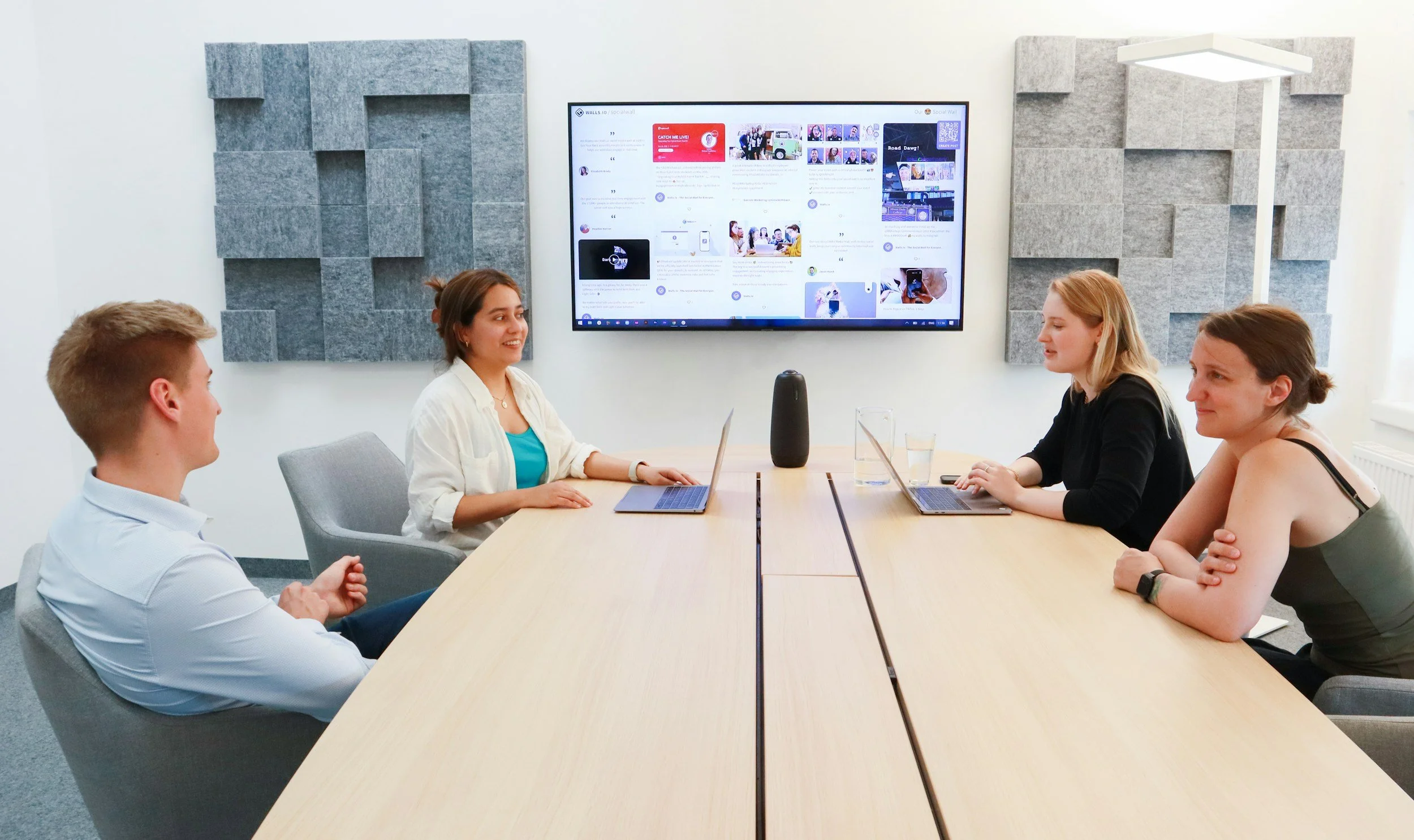 A group of five people seated around a conference table in a meeting room, with a large screen displaying social media content behind them.