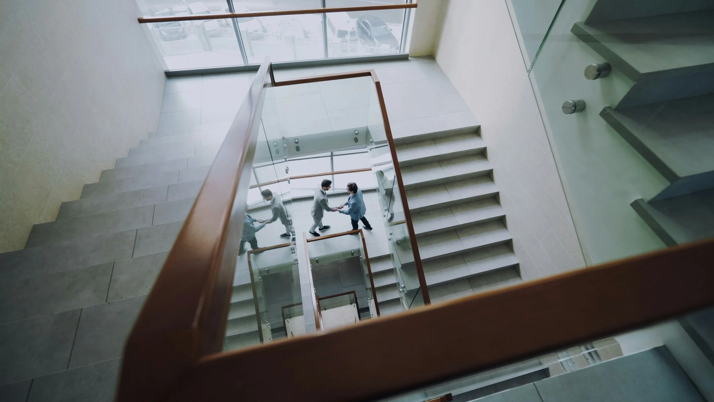 View of a modern multi-story building interior taken from the top of a staircase, showing two men shaking hands. The staircase has glass sides and wooden handrails, with large windows letting in natural light and a view of the outside.