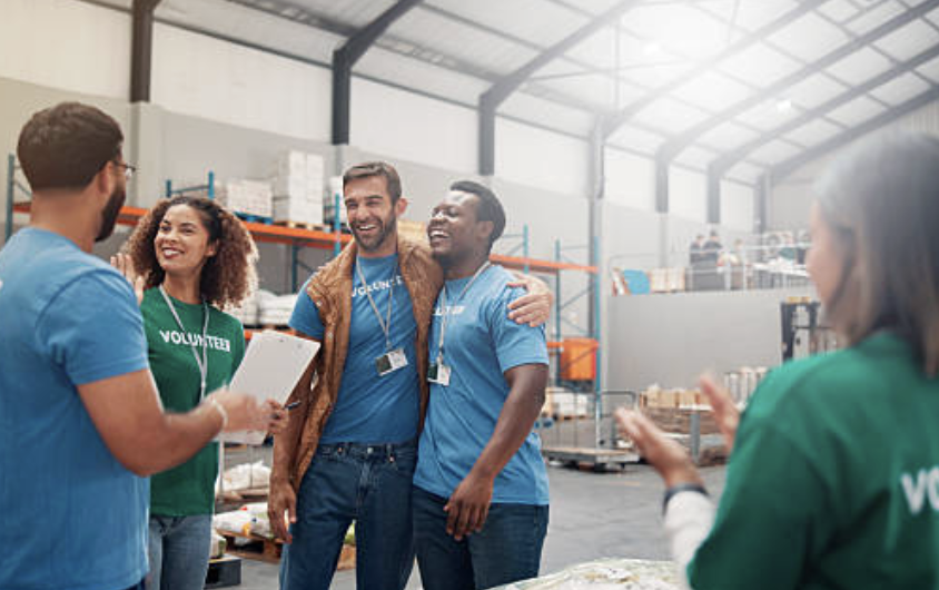 Group of volunteers smiling and talking in a warehouse setting, wearing volunteer shirts.