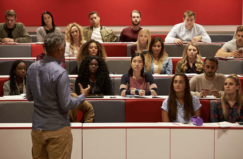 A man is speaking to a group of students in a classroom with tiered seating, with a red wall behind them.
