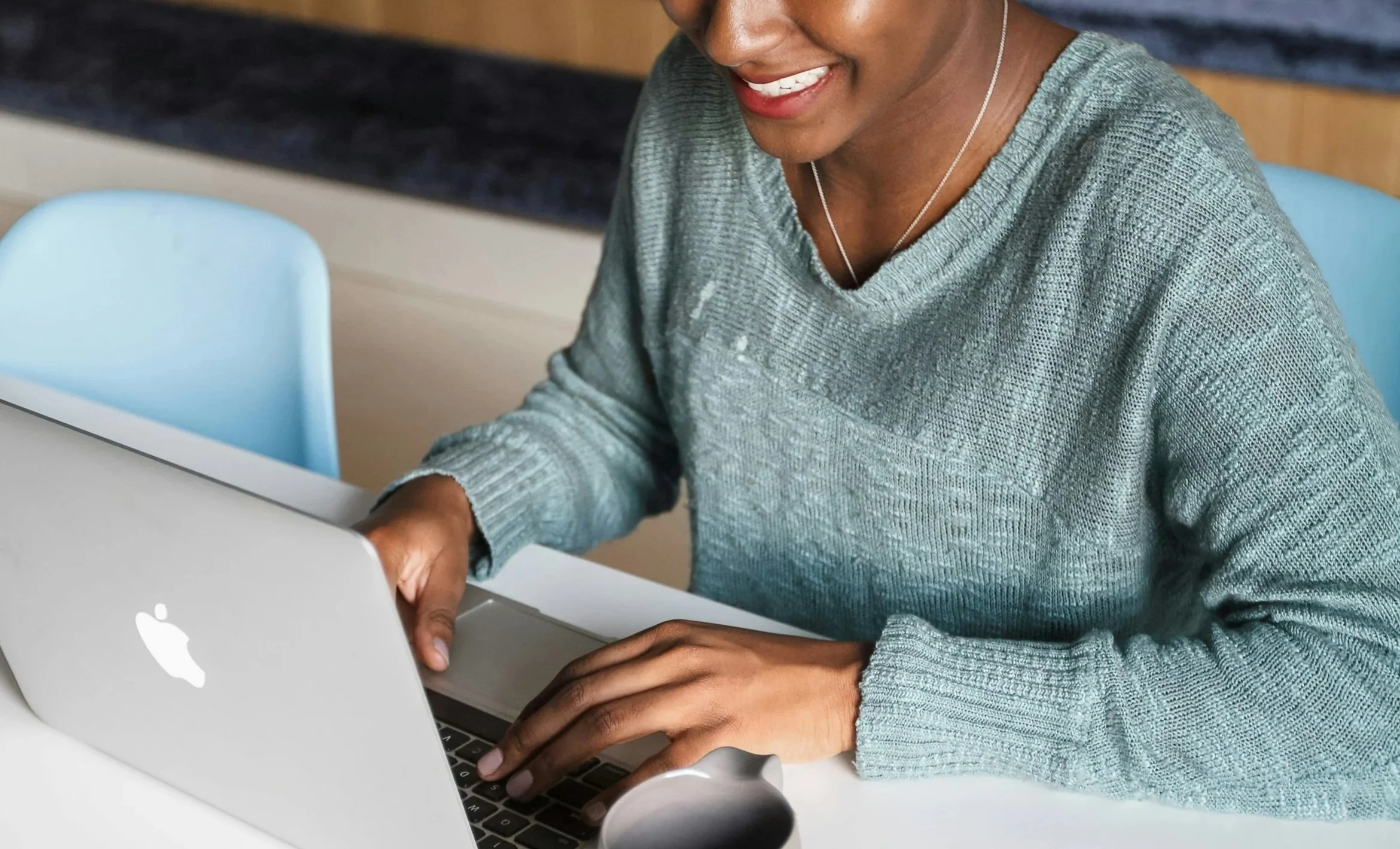 A woman smiling while working on a silver MacBook in a casual setting, wearing a green sweater and a silver necklace.