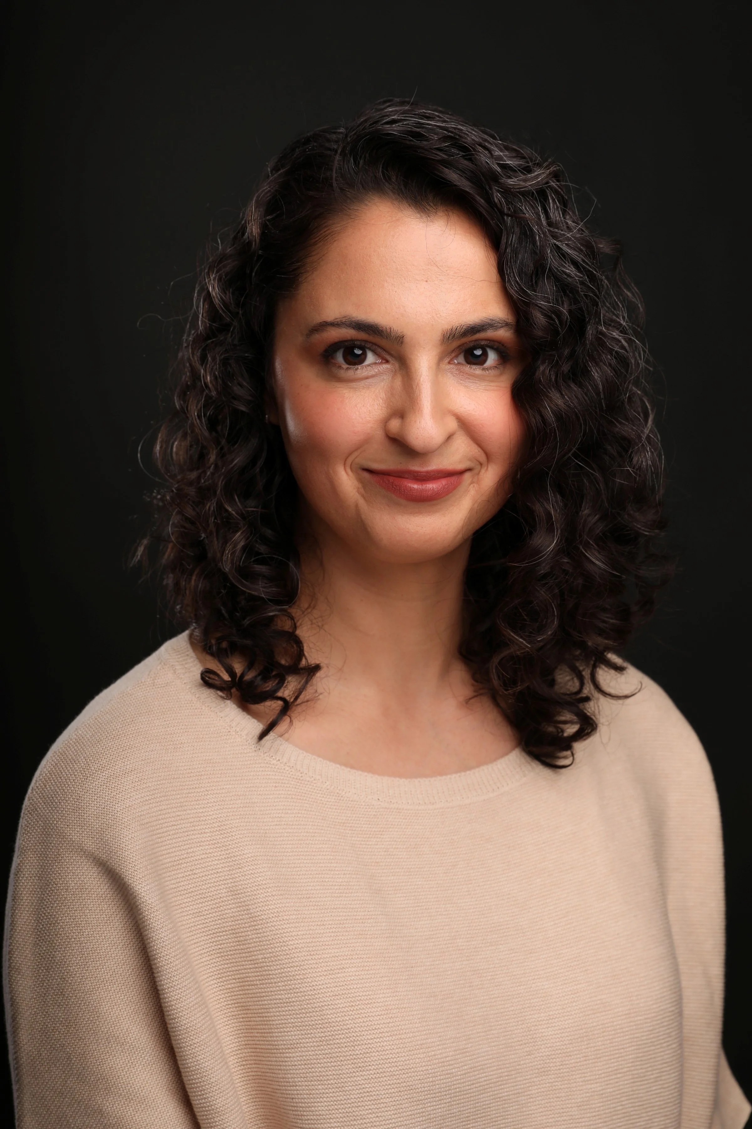 A woman with curly dark hair, wearing a beige sweater, smiling softly, against a black background.
