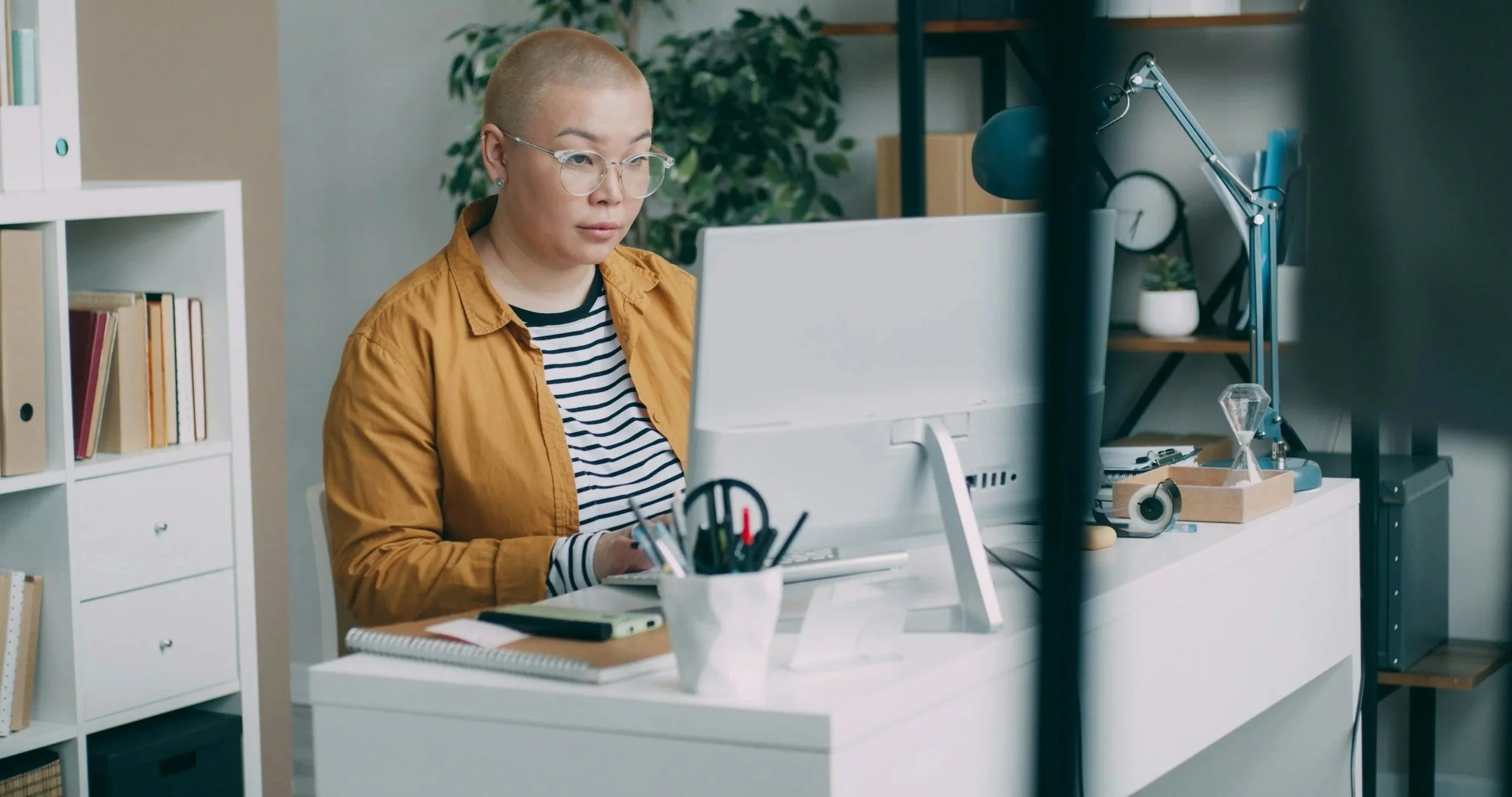A woman with glasses and short hair working at her desk in a home office, surrounded by books and office supplies.