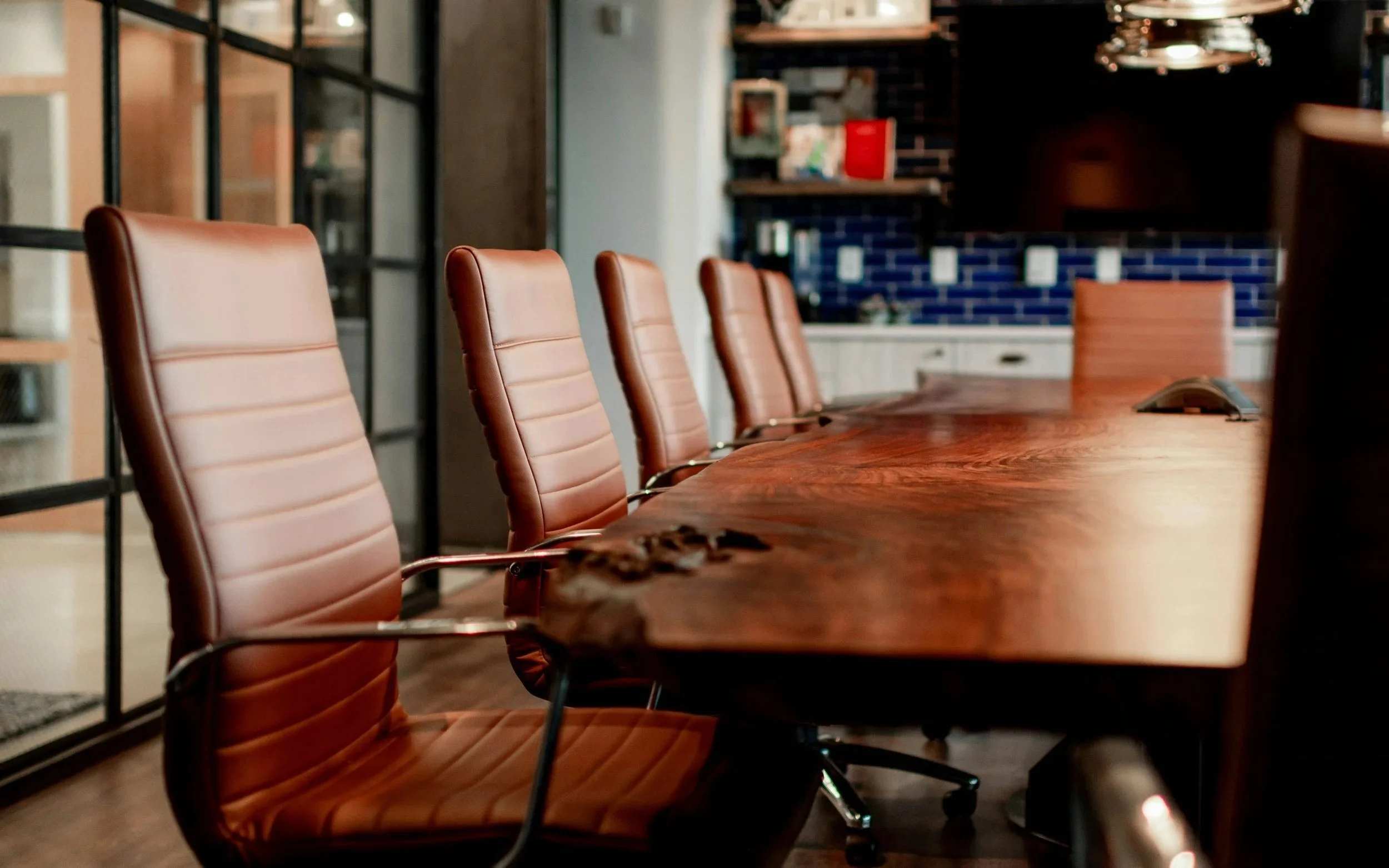 A long wooden conference table with several tan leather chairs arranged around it in a modern office or meeting room. In the background, there are shelves, a brick wall, and a kitchen area.