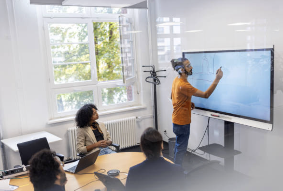 Man giving a presentation on a digital whiteboard to a group in a meeting room with large windows.
