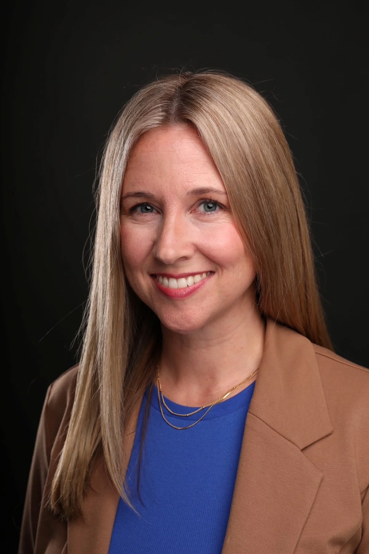 A woman with long blonde hair, blue eyes, and fair skin, smiling, wearing a blue top, layered gold necklaces, and a tan blazer against a black background.