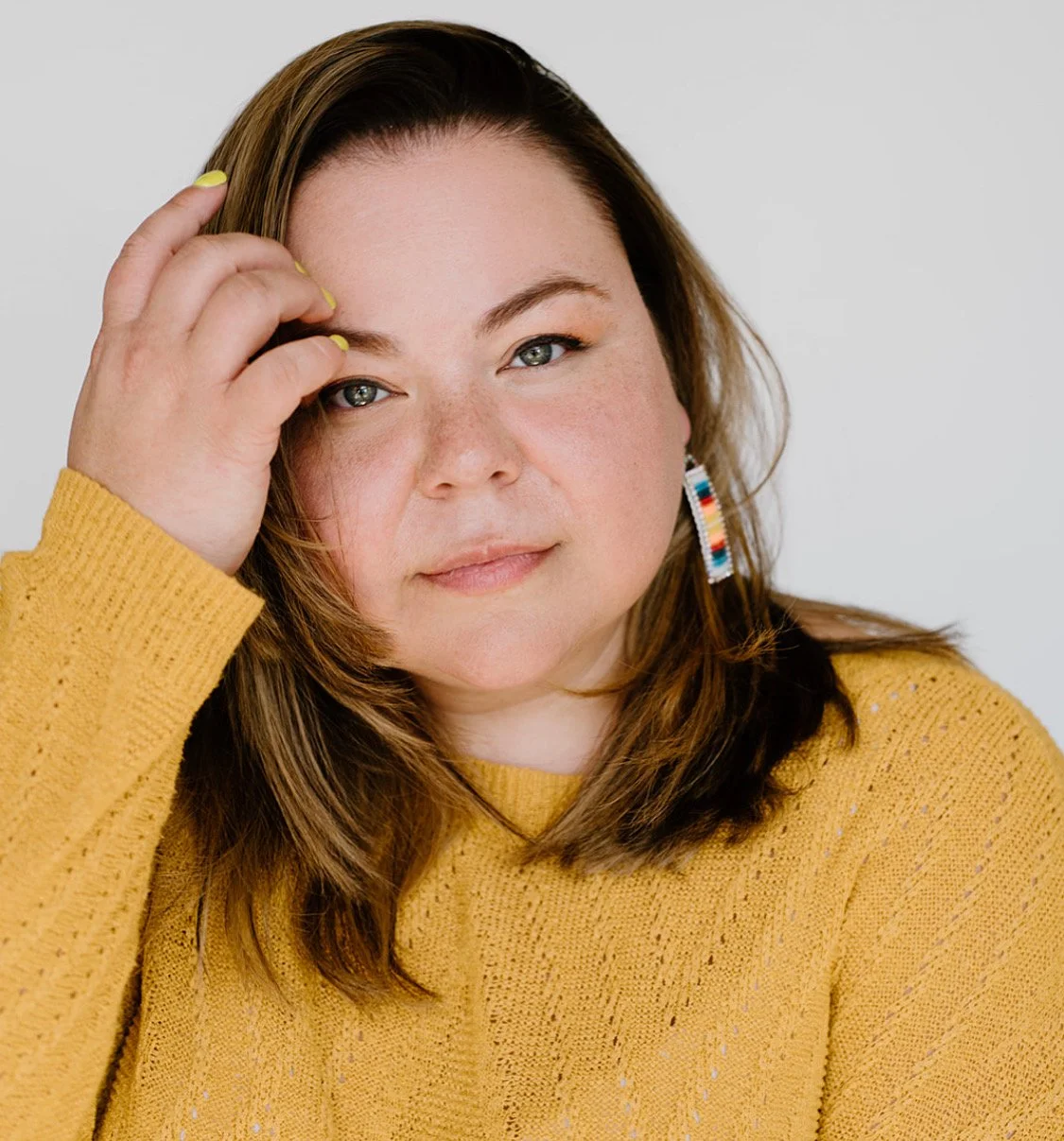 A woman with brown hair, blue eyes, and freckles, wearing a yellow sweater and colorful beaded earrings, touching her forehead with her hand, looking directly at the camera.
