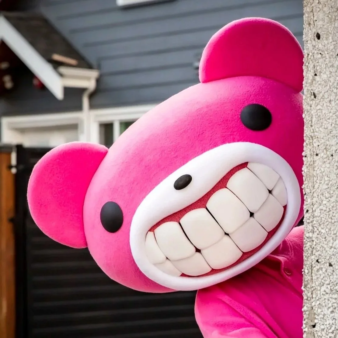 A pink cartoon character with oversized head, big teeth, and black eyes peeking around a wall.