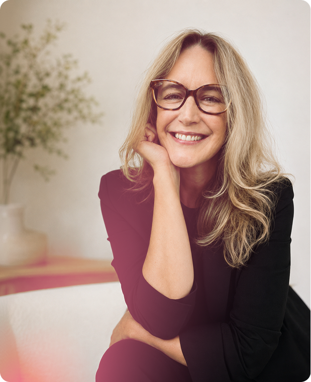 A smiling woman with long wavy blonde hair wearing glasses and a black top, sitting indoors with a blurred green plant in the background.