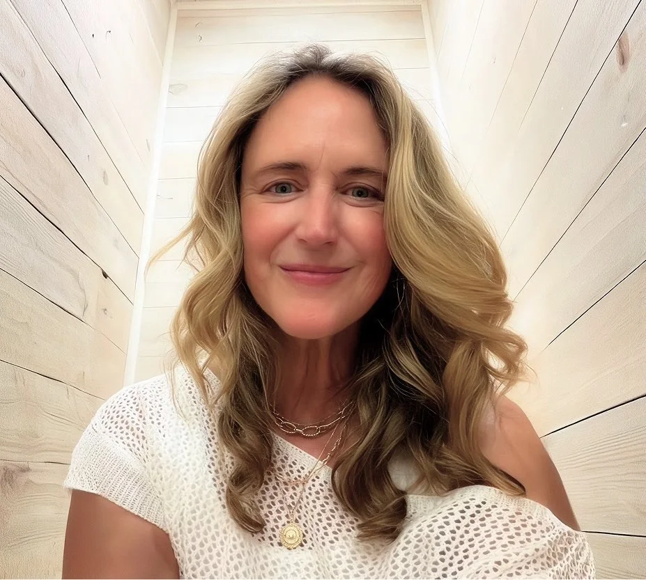 A smiling woman with wavy blonde hair taking a selfie in a narrow, light-colored wooden hallway.