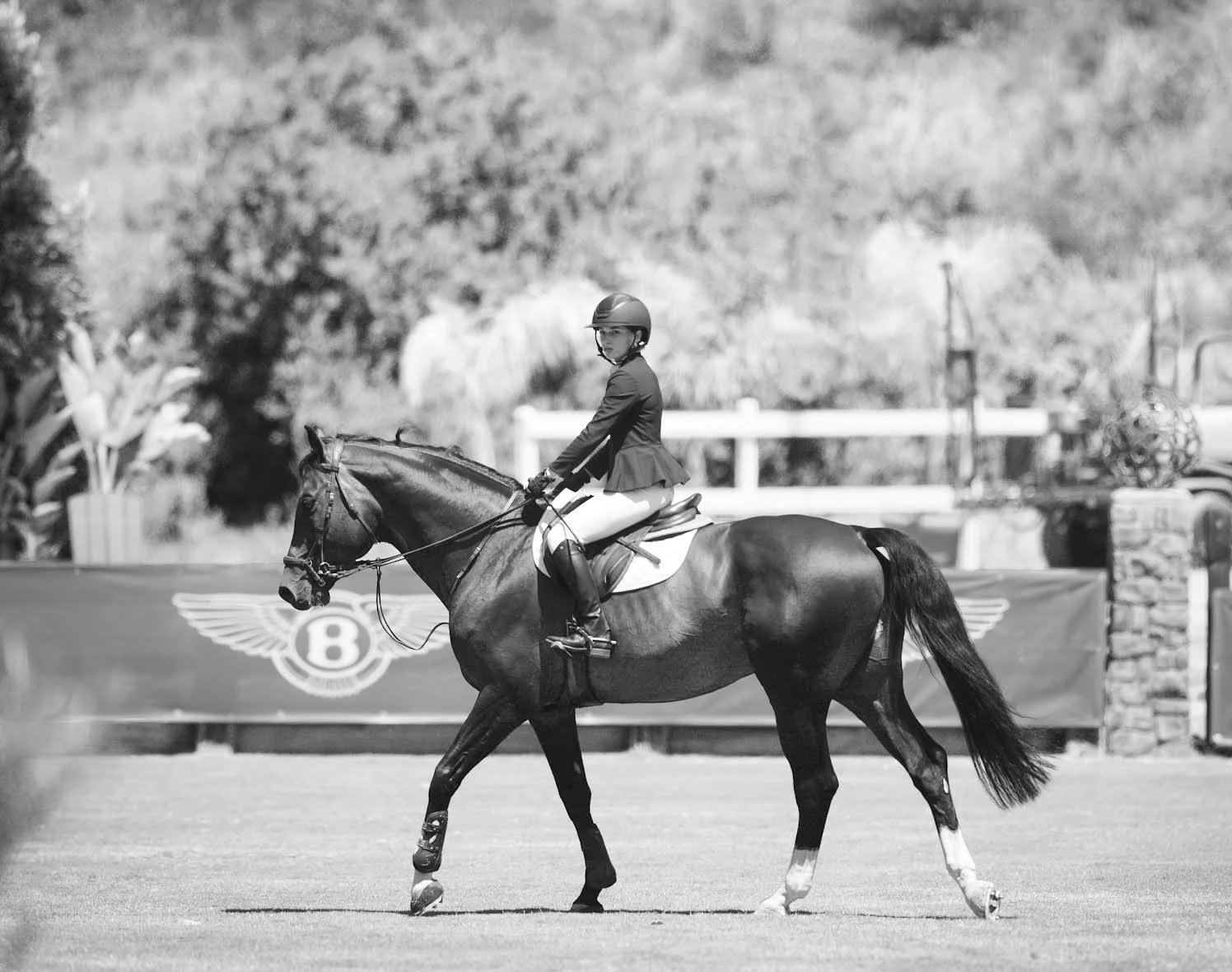 A woman riding a horse in an equestrian arena with a Bentley logo banner in the background, in black and white.