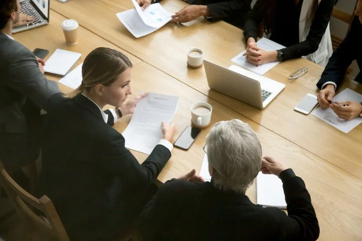 Business meeting with people sitting around a wooden table, reviewing documents and using laptops and smartphones.