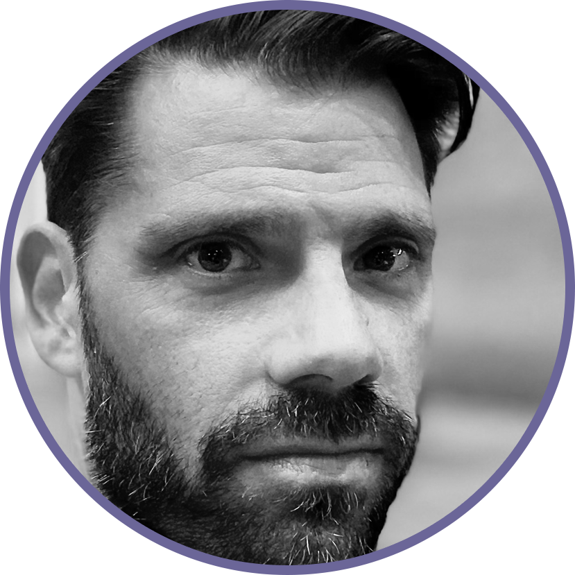 Close-up of a man with dark hair and a beard, looking directly at the camera in black and white.
