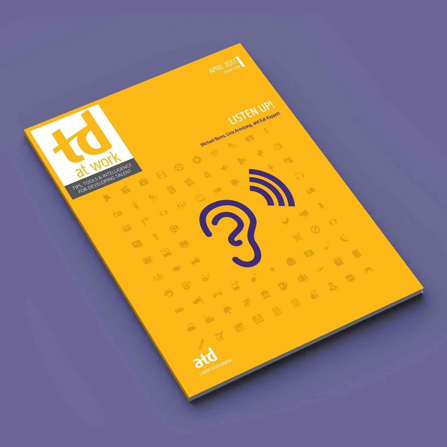 Magazine titled 'at work' with a yellow cover, featuring a large blue ear with Wi-Fi signals, and a background pattern of small icons related to communication and work, placed on a purple surface.