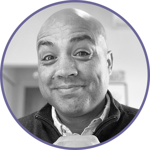 Black and white photo of a man with a shaved head smiling, wearing a collared shirt and a zip-up jacket, in an indoor setting.