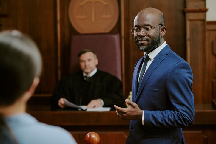 Man in blue suit speaking in a courtroom with judge and person in the foreground.