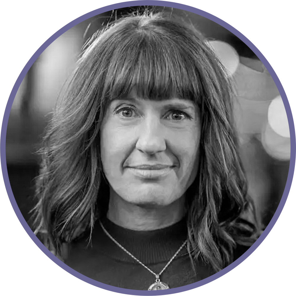 Black and white photo of a woman with wavy hair and bangs, wearing a necklace with a pendant, smiling slightly.