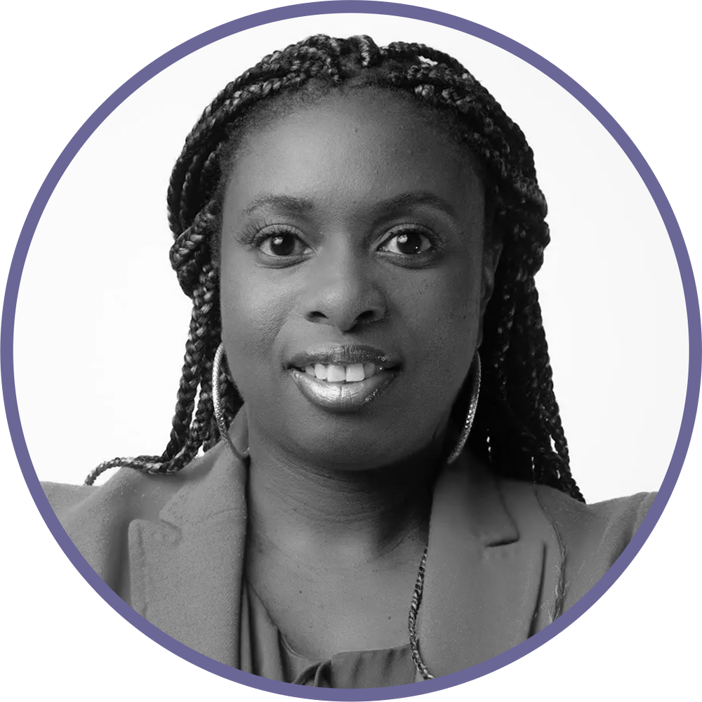 Black and white portrait of a woman with braided hair, wearing hoop earrings and a blazer, smiling at the camera.