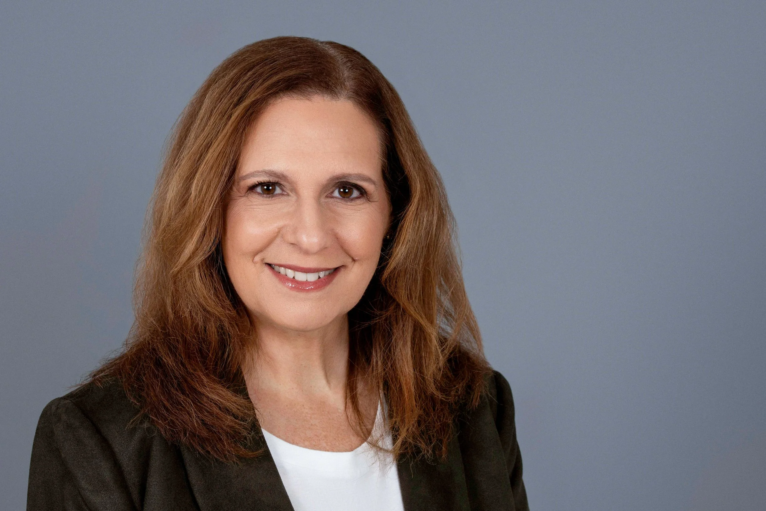 A woman with shoulder-length reddish-brown hair, smiling at the camera, wearing a black blazer and white top, against a plain blue background.