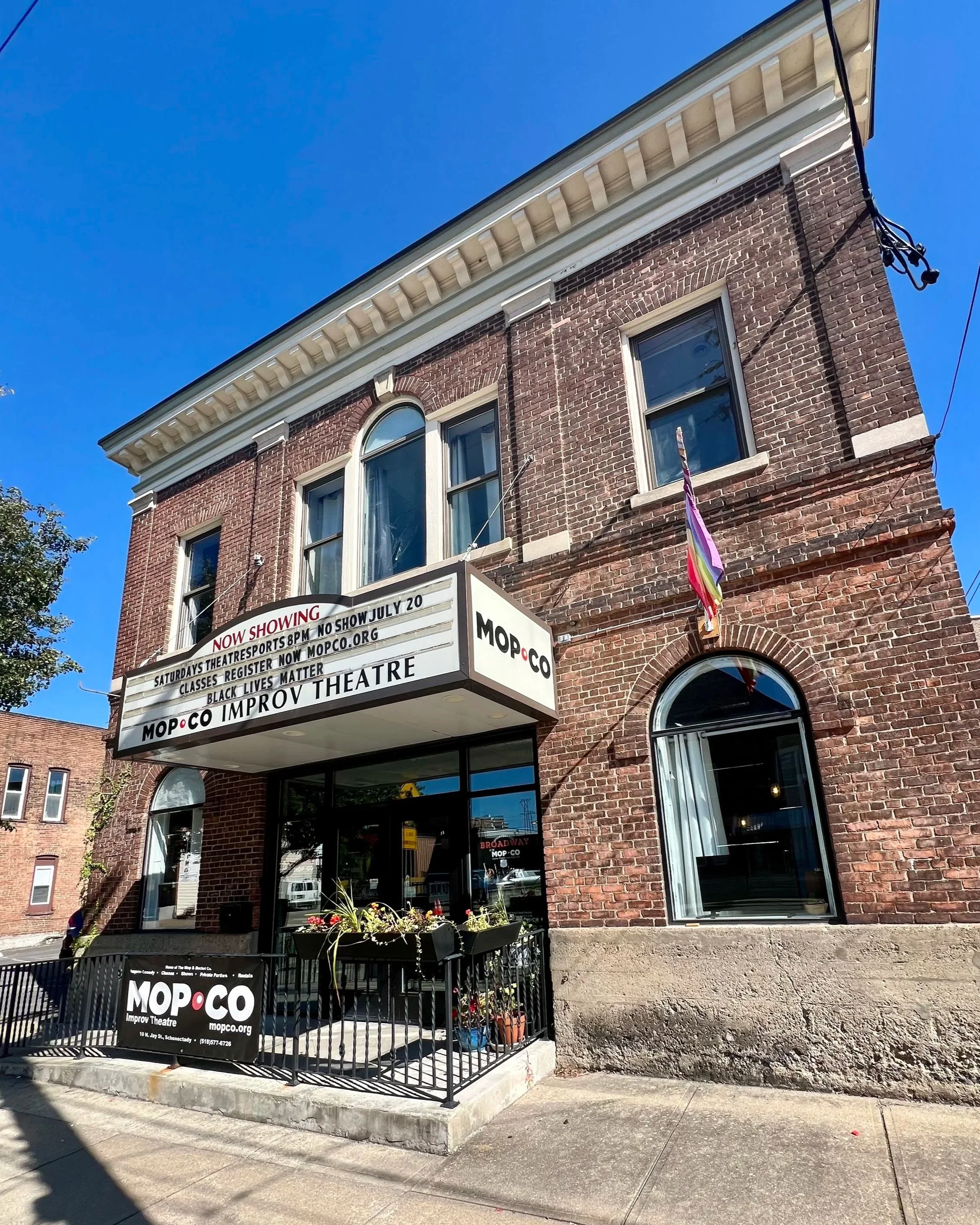 Brick theater building with marquee sign advertising improv shows, flags on the side, and a sidewalk in front.
