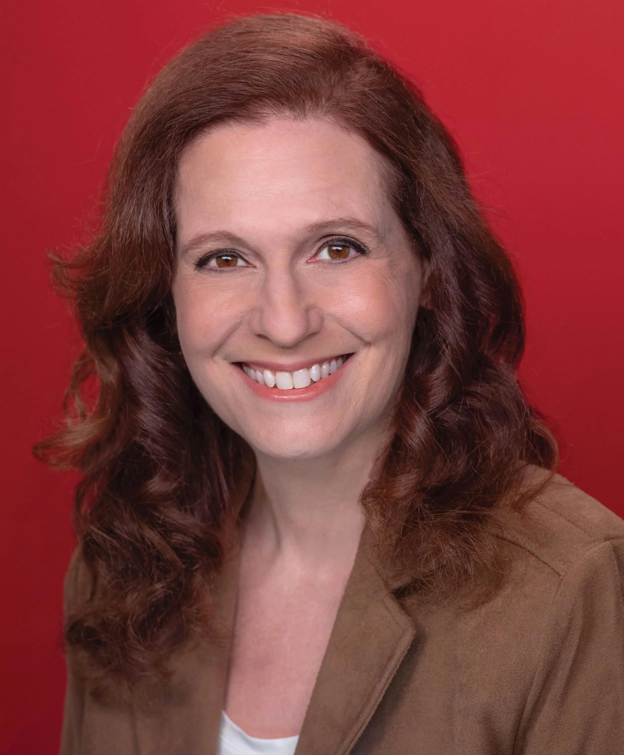 Portrait of a smiling woman with wavy brown hair, wearing a brown jacket against a red background.
