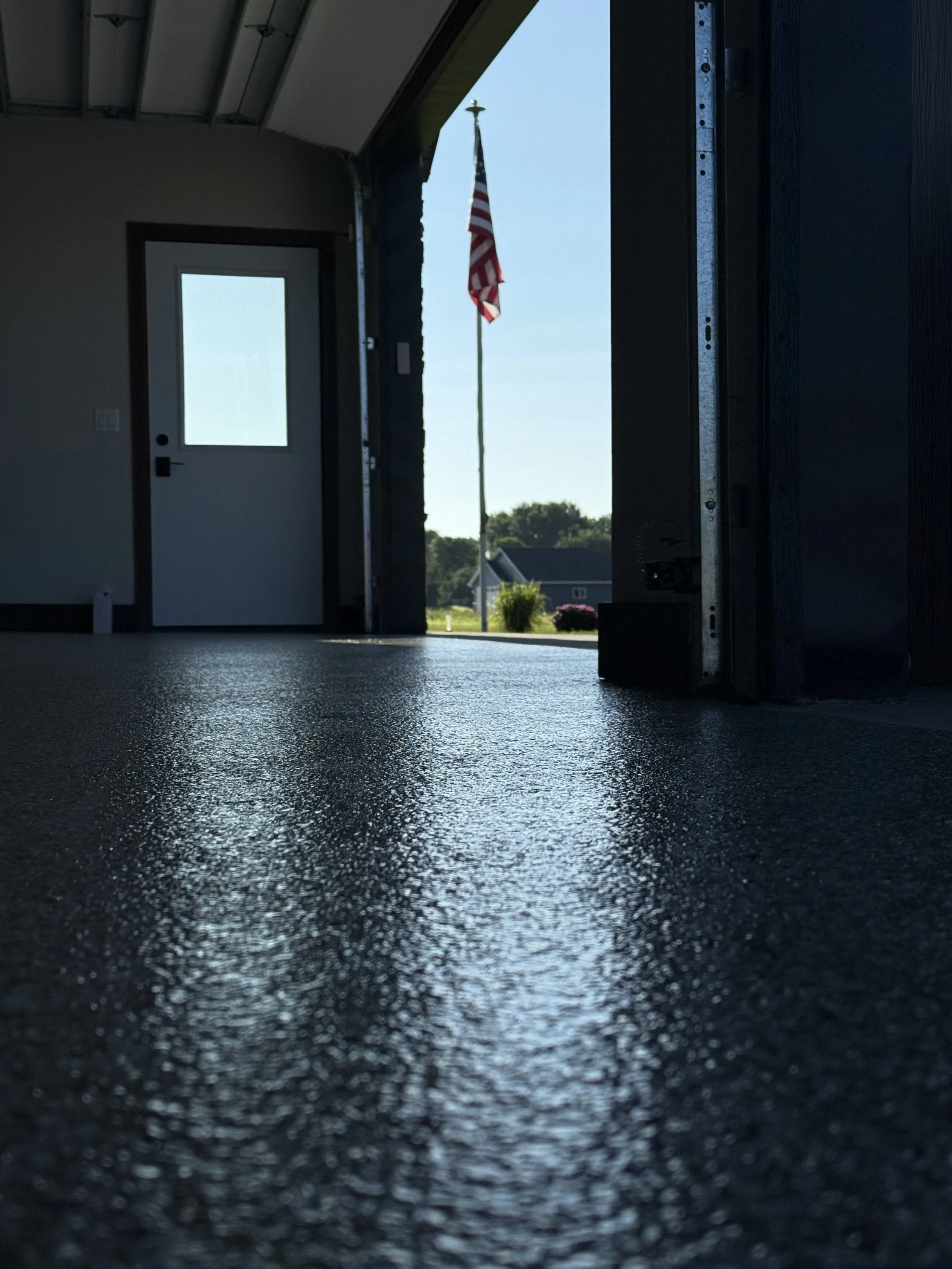 Looking inside a garage with an American flag outside, providing a view of the yard and neighboring houses in the background during daytime.
