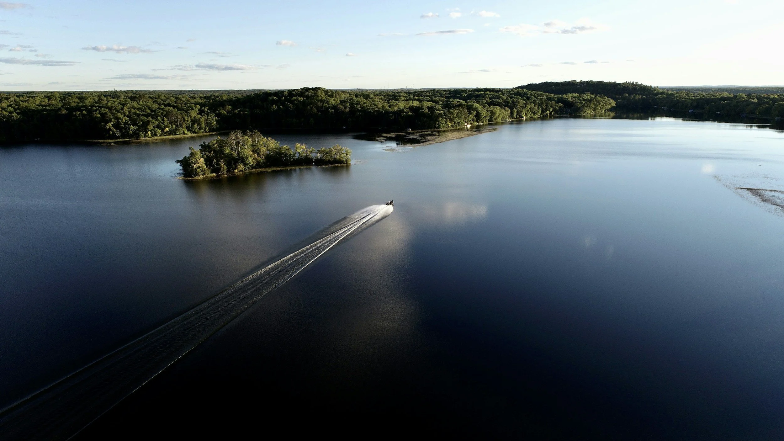 Aerial view of a boat cruising on a large calm lake with small islands and forested shoreline.