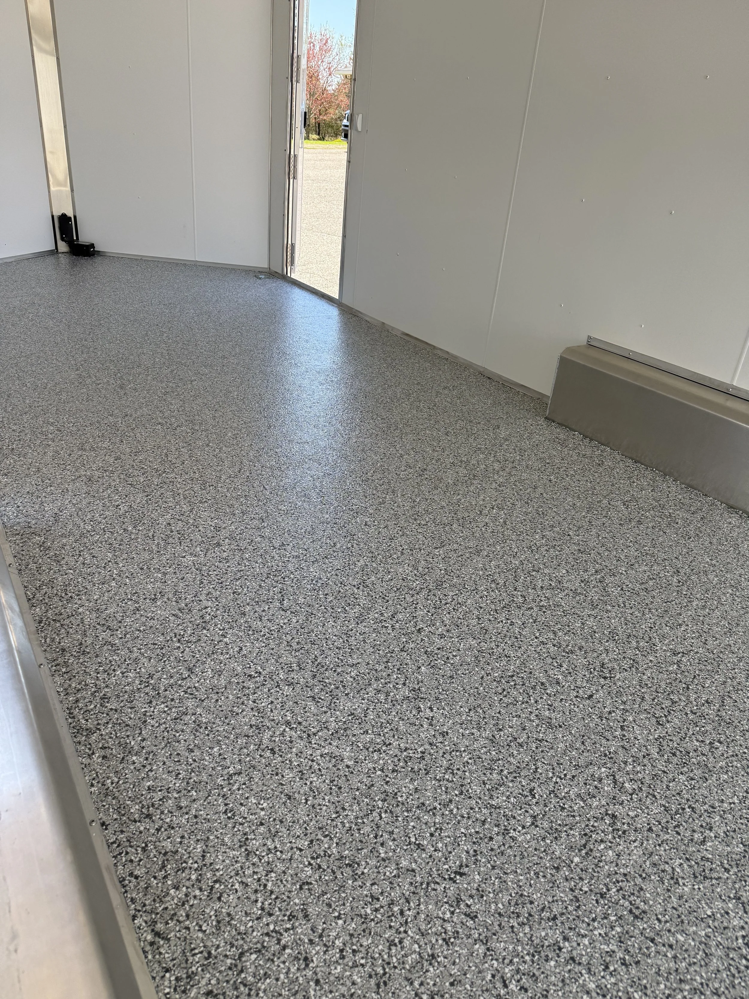 View of the interior of an empty mobile or trailer with speckled gray flooring, white walls, and an open door showing a parking lot and trees outside.