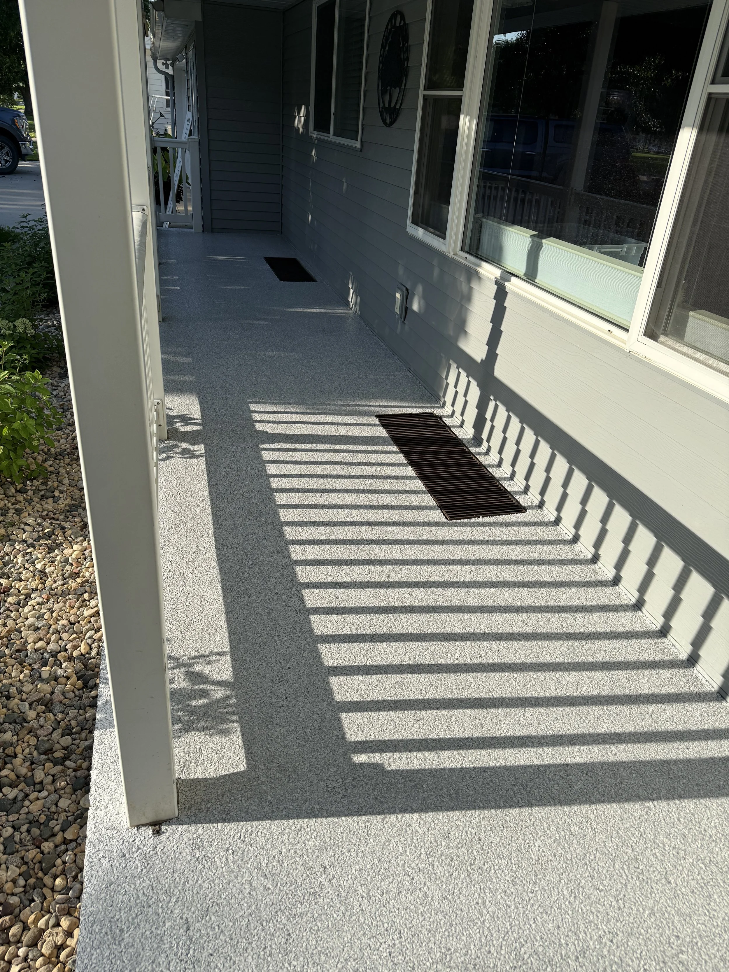 A porch with a textured white floor, gray house exterior, windows, and two vents or drain grates. Shadows from the railing create striped patterns on the porch