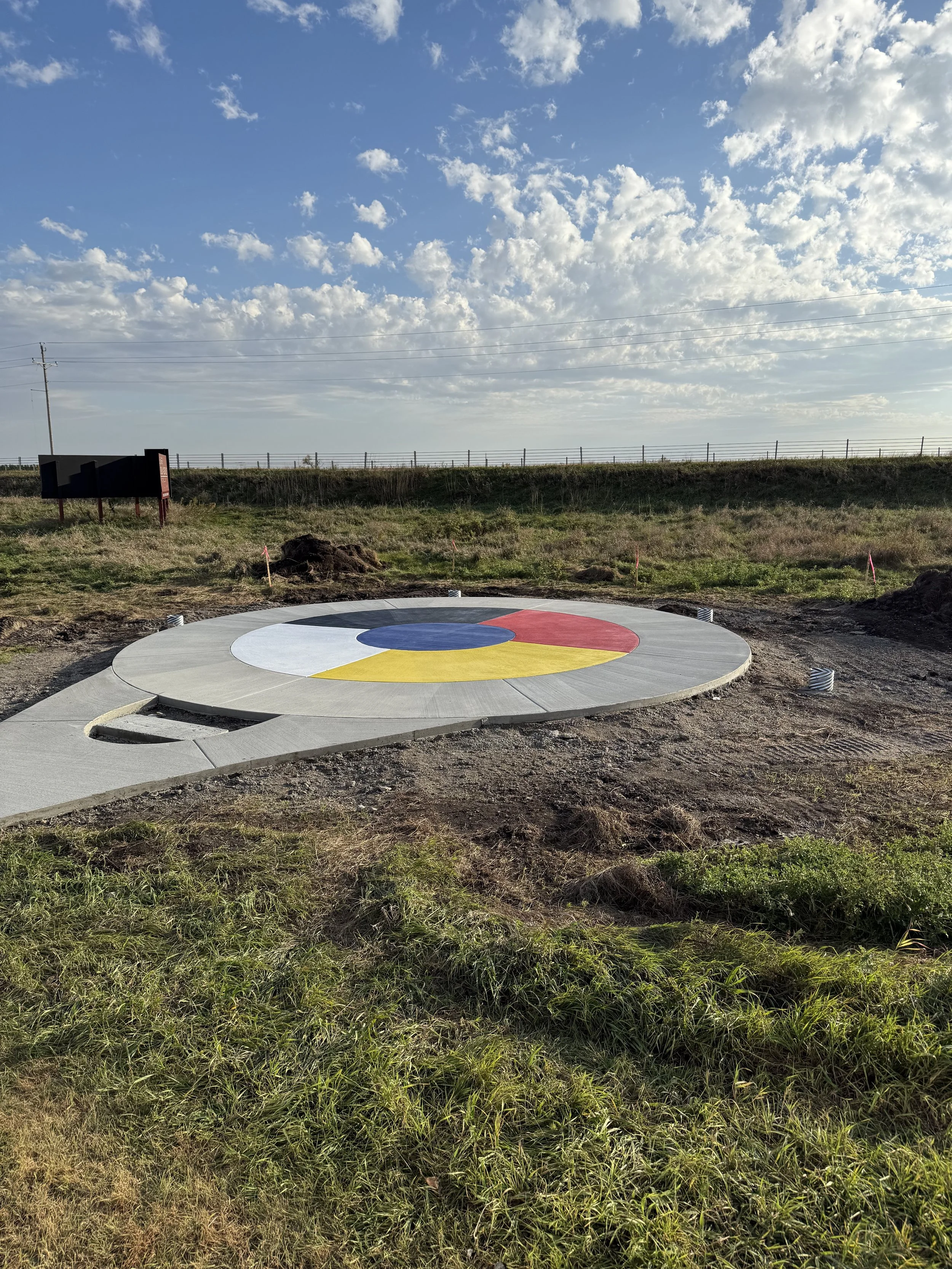 A freshly painted curling stone target with red, yellow, blue, black, and white concentric rings on a concrete pad outdoors, with a cloudy sky and grassy field in the background.