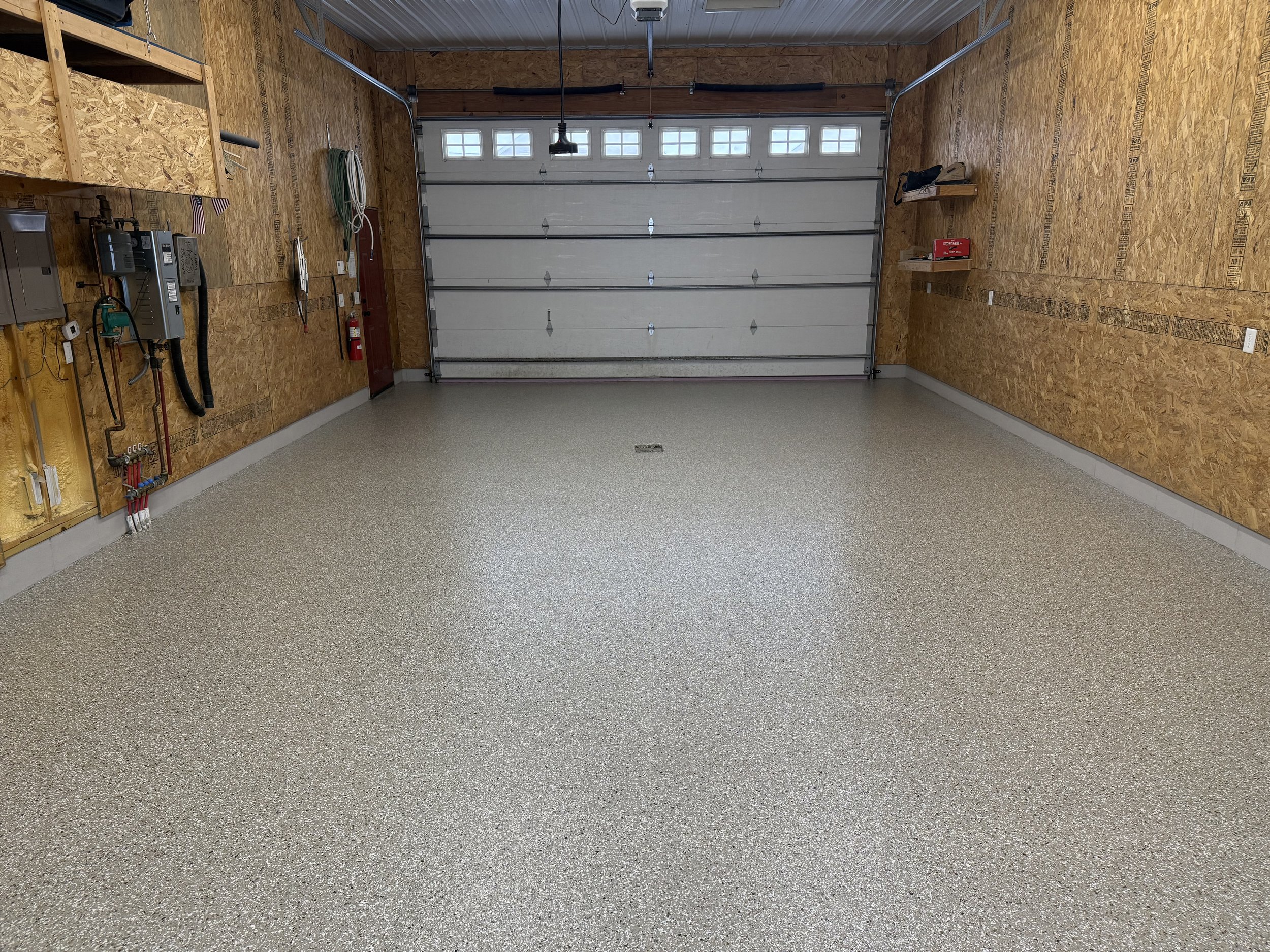 Empty garage with a textured, speckled floor, unfinished wood walls, a large garage door, and shelves with miscellaneous items.