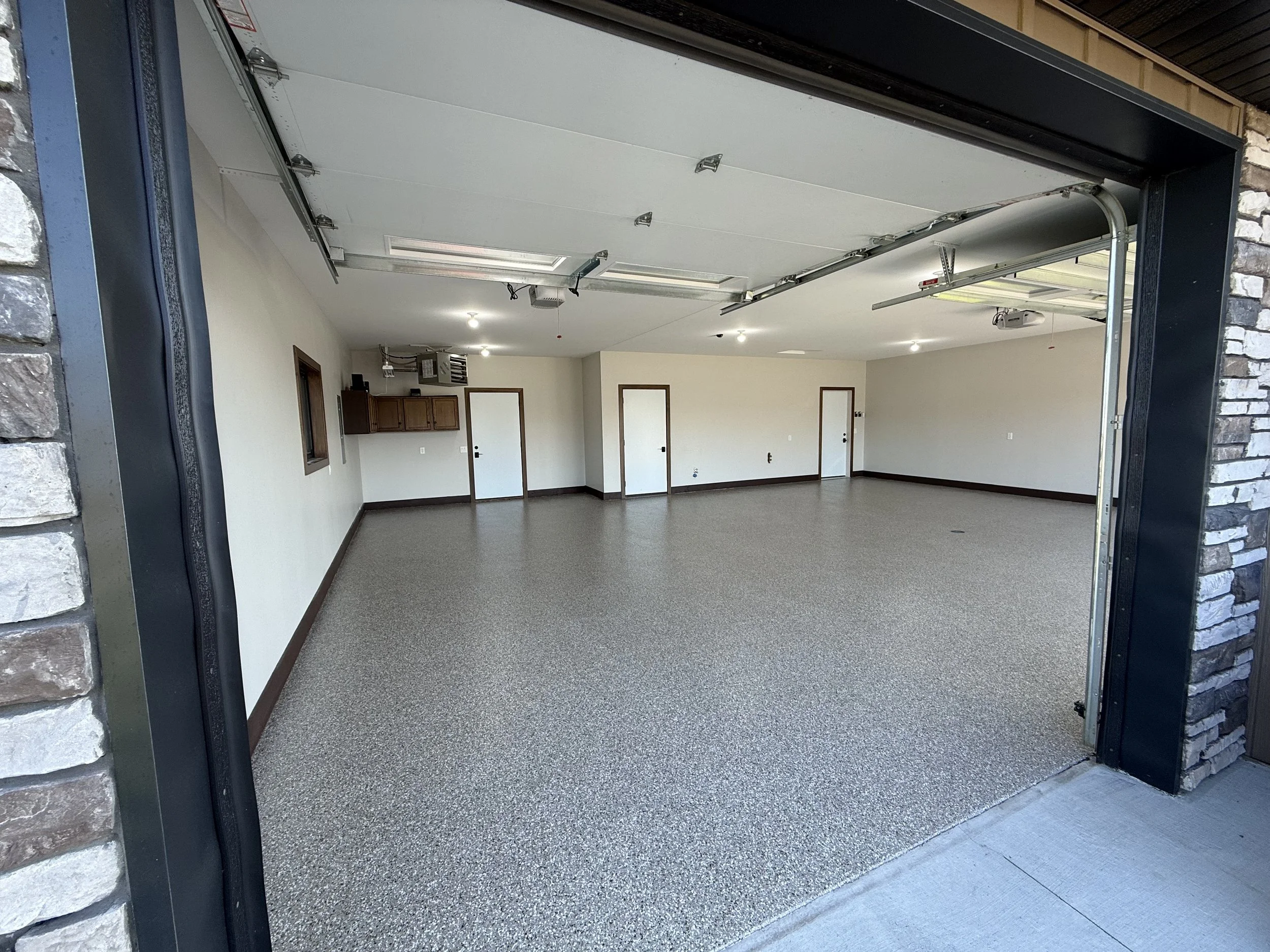 Empty residential garage with speckled epoxy floor, white walls, wood baseboards, and three doors on the back wall. The garage door is open, exposing the interior, with a small window and some cabinets visible on the left side.