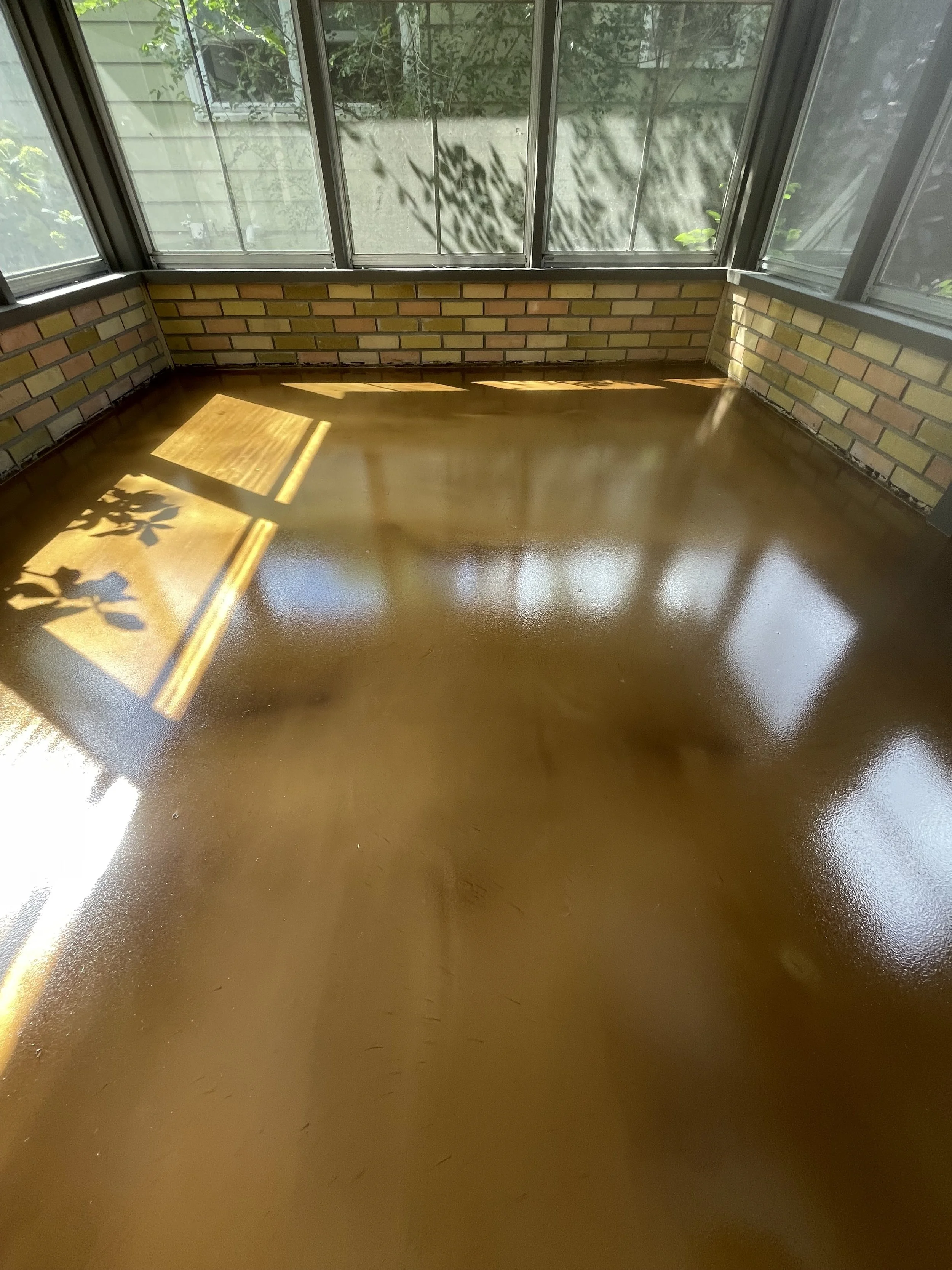 Sunlight coming through window casts shadows onto a shiny, polished wooden floor in a sunroom with brick walls and large windows.