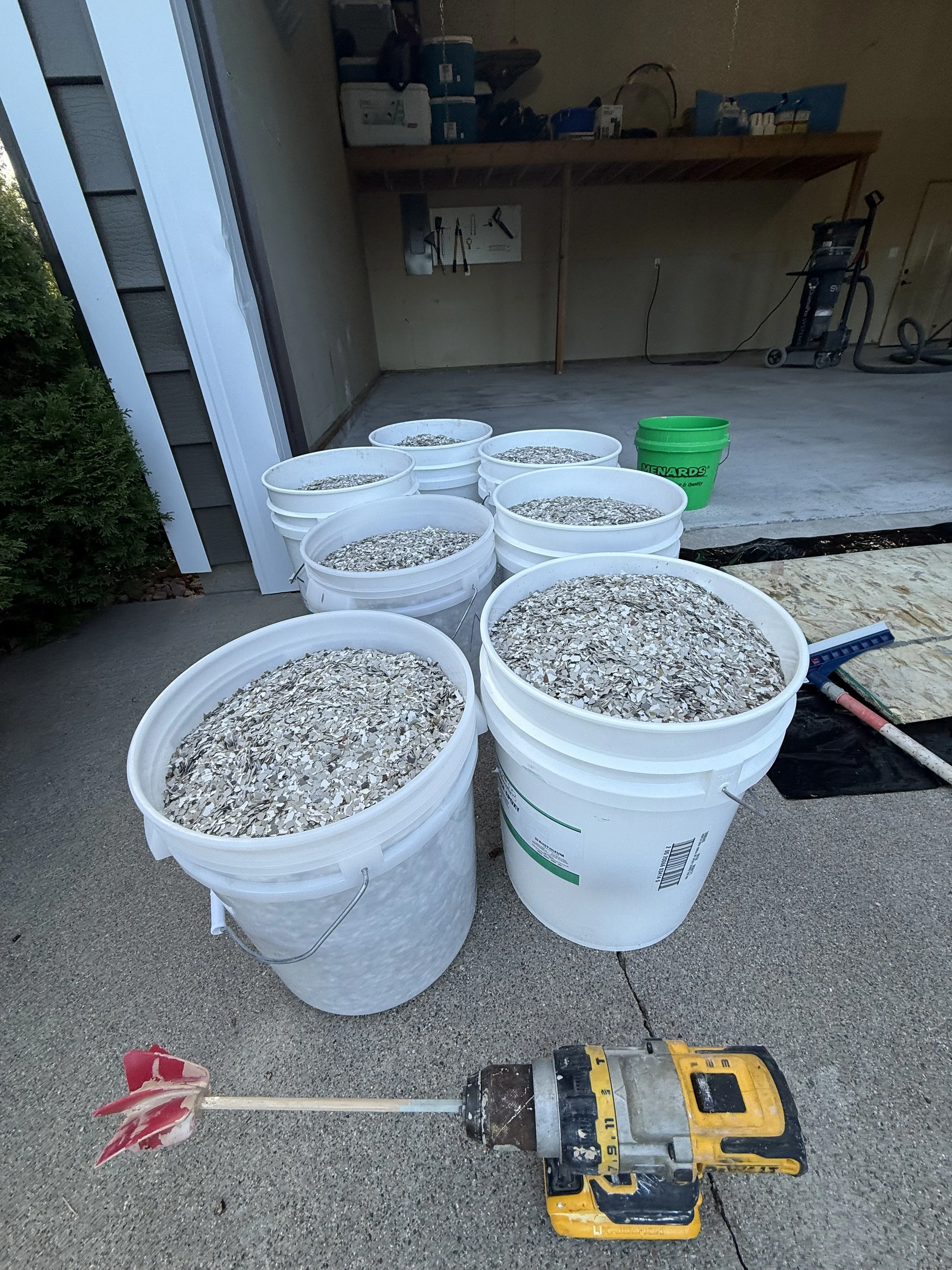Multiple white buckets filled with fleck, placed outdoors on a concrete surface near a garage.