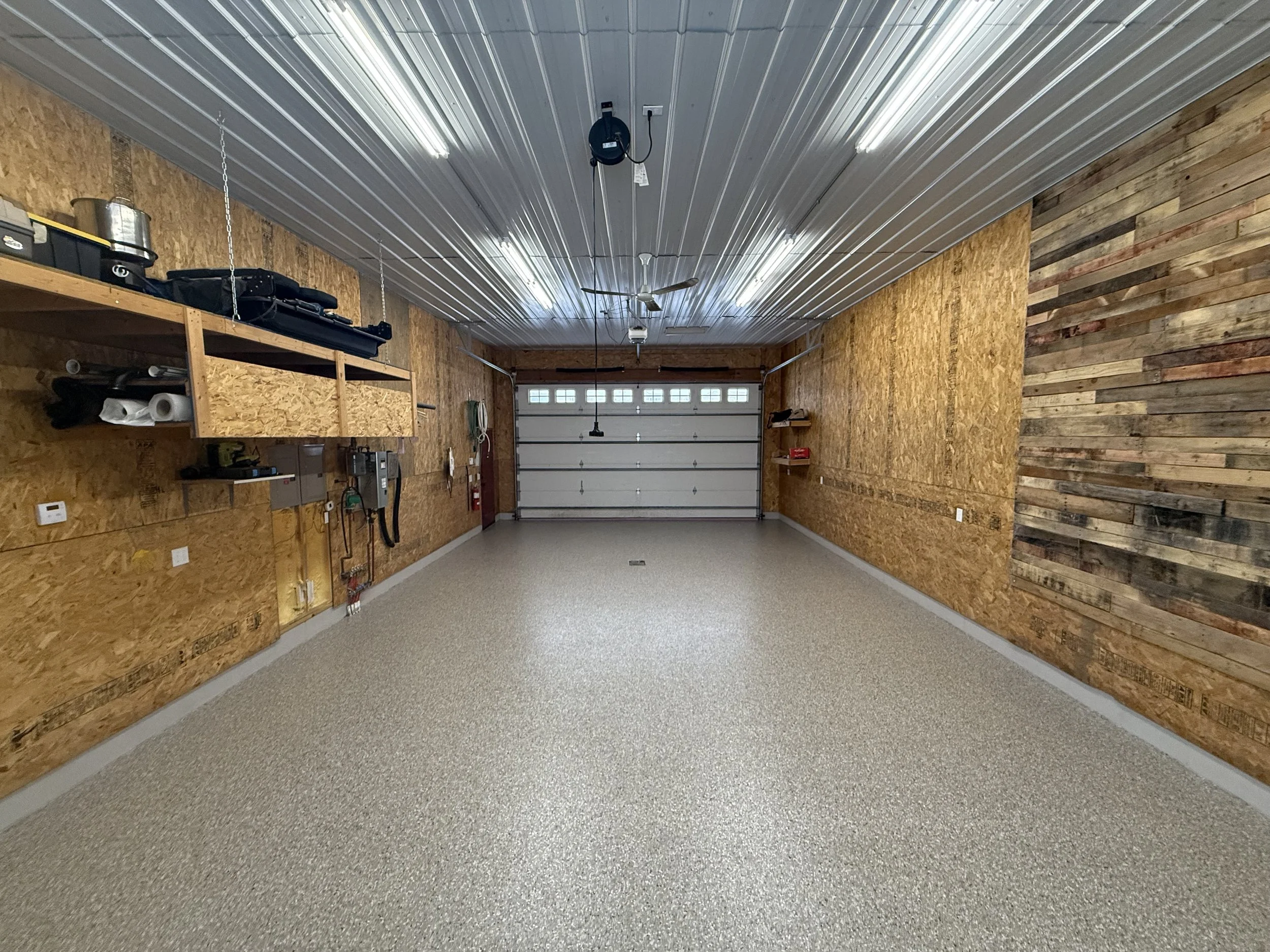 Empty garage with wooden and metal wall panels, overhead fluorescent lighting, a garage door at the end, and wall-mounted shelves with tools and supplies.