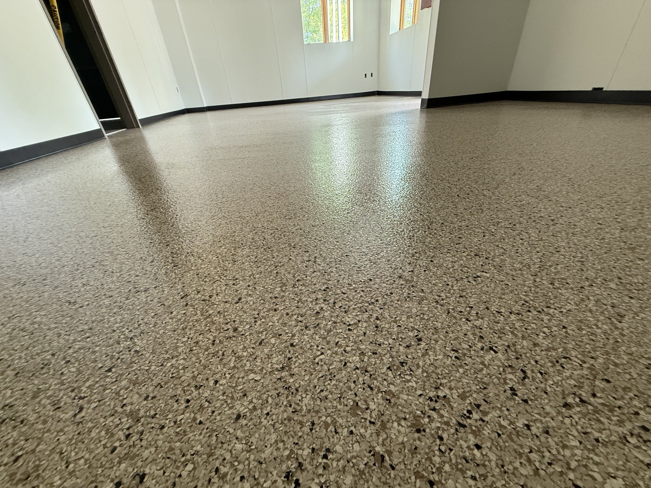 Empty room with speckled tan flooring, white walls, black trim, and windows showing greenery outside.